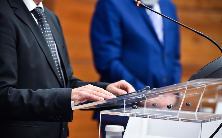 man at podium in suite speaks on a microphone